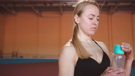 atleta rubia bebiendo agua de una botella en una instalación deportiva cubierta