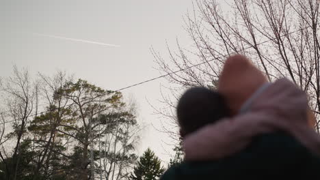 a man in a green jacket carries his daughter, dressed in a pink jacket and matching beanie, with her head gently resting on his as they both look up at the sky. the father points towards the sky