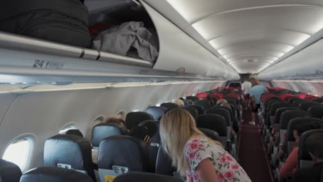 blonde woman reaches up to open overhead bin storage and stuffs large backpack to store