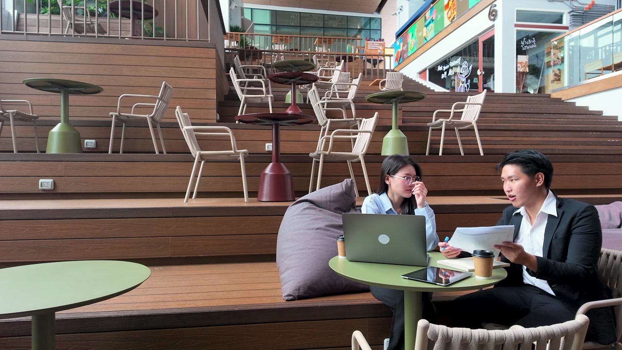 Thai professionals discuss paperwork and laptop data at a modern outdoor office in Bangkok.