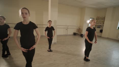 a group of young ballet students in black dancewear practicing positions in a spacious ballet studio with wooden flooring and wall-mounted barres. focused expressions and synchronized movements.