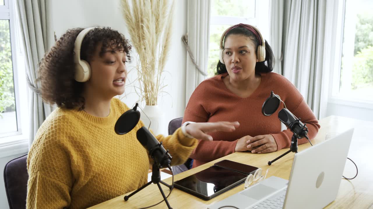 Co-conduttrici di varie etnie che parlano e gesticolano a casa con microfoni, usando un laptop per un podcast.