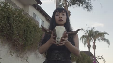 pretty woman in steam-punk clothing holding a goat head with red eyes in an alley