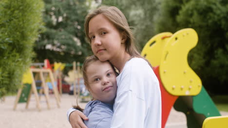 Close-up-view-of-a-little-girl-with-down-syndrome-hugging-another-girl-while-they-looking-at-camera-in-the-park-on-a-windy-day