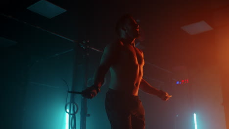 Muscular-man-with-bare-torso-is-jumping-using-a-skipping-rope.-Male-boxer-with-skipping-rope.-Fighter-doing-jump-rope-exercises-in-colorful-light-with-smoke-in-slow-motion.