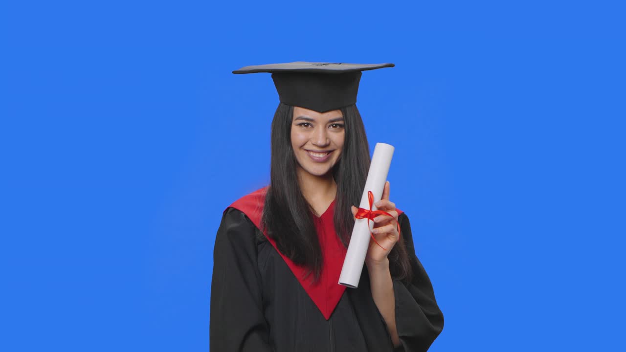 retrato de una estudiante en traje de graduación de gorra y túnica, sosteniendo un diploma y haciendo un gesto de roca. mujer joven posando en el estudio con fondo de pantalla azul. primer plano. cámara lenta lista 59.94fps