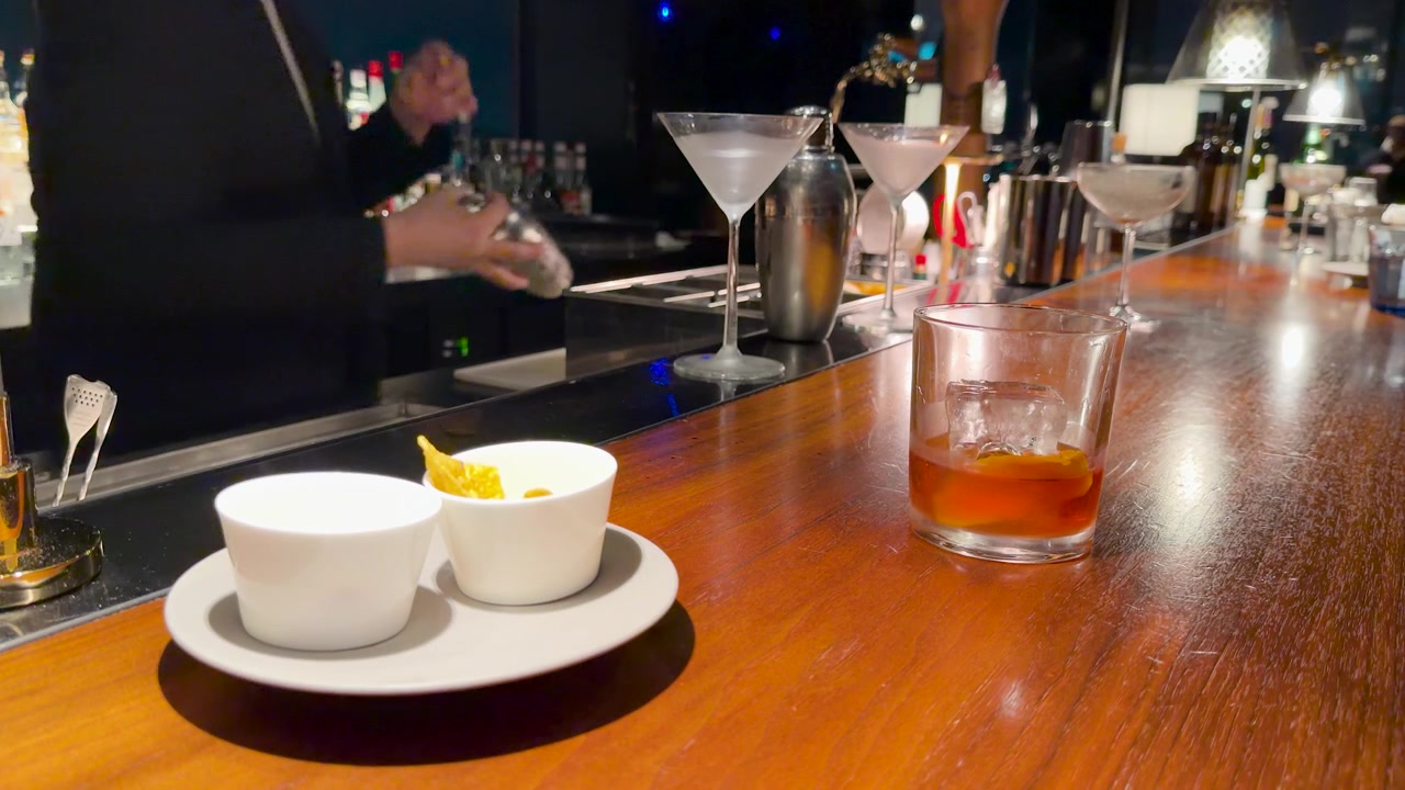 Một bartender mặc vest đen đang lắc cocktail phía sau quầy bar gỗ bóng loáng.