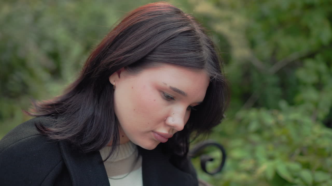 공원 벤치에서 책을 읽는 우아한 여성의 중간 측면 뷰, 손으로 귀 옆의 머리카락을 정리하고, 가을빛이 있는 부드럽게 흐릿한 숲 배경, 차분하고 집중된 분위기, 조용한 자연의 순간