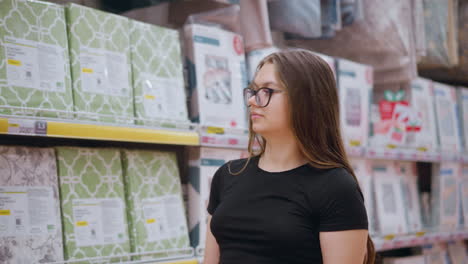 student in black top and glasses walking through supermarket aisle looking at household goods on shelves, pausing to examine products before turning attention to something else in background