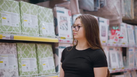estudiante con camiseta negra y gafas caminando por el pasillo del supermercado mirando los artículos para el hogar en los estantes, deteniéndose para examinar los productos antes de dirigir la atención a algo más en el fondo