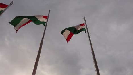 Three-Hungarian-flags-on-flagpoles-blowing-in-the-wind-with-storm-clouds-in-the-background---Slow-motion-dolly-shot-looking-upwards