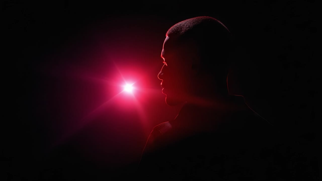 Une silhouette se dresse sur un fond de lumière rouge intense, créant un contraste saisissant entre l’obscurité et l’illumination, évoquant le mystère et l’intrigue.