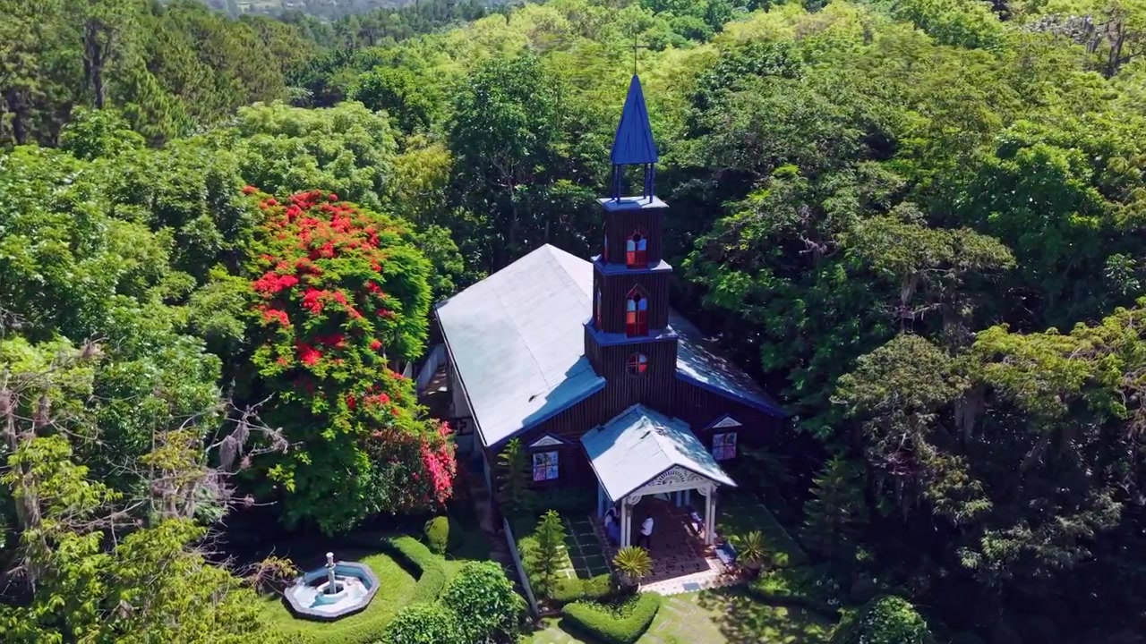 La torre de la Capilla La Milagrosa se eleva a través del dosel tropical en San José de las Matas, República Dominicana; una aguja azul ornamentada corona las vidrieras sobre la entrada del jardín. Vista aérea de dron cenital