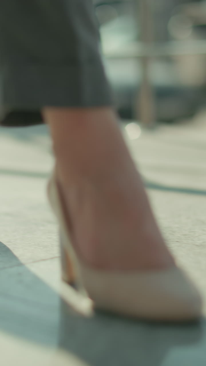 Close up leg view of woman in heels walking gracefully beside iron railing with parked cars in background, sunlight casting soft shadows on pavement as she moves confidently