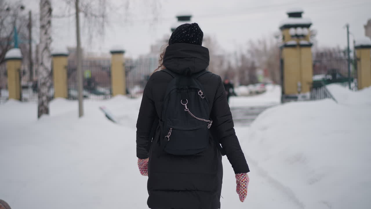 Mujer caminando por un parque nevado en la ciudad, vista posterior, con abrigo negro, mochila, gorro, guantes, bancos, entorno urbano, estación fría, exteriores, soledad, ambiente gélido, camino helado, entorno tranquilo