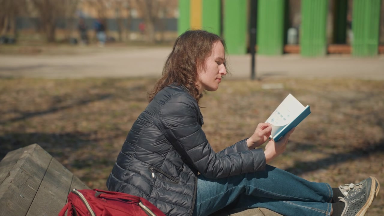 Woman reads book on wooden bench in park with red backpack sunlight and greenery background reading outdoor sitting daytime nature text closeup relaxed