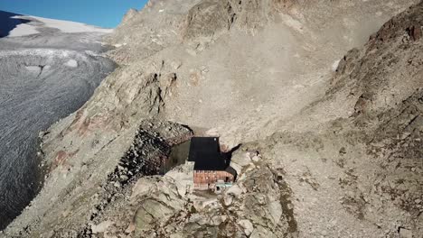 Drone-view-of-a-wooden-cabin-in-the-swiss-alps-on-a-steep-rocky-mountain-next-to-a-large-glacier