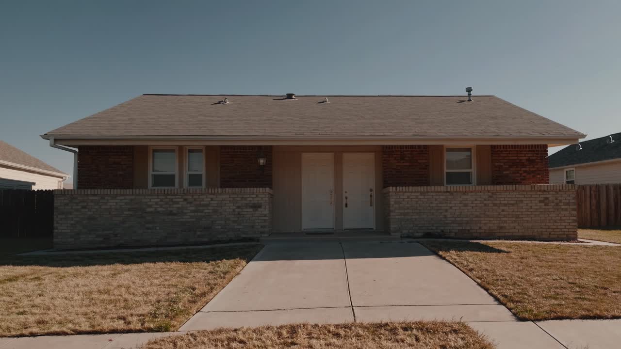 Small duplex house featuring a brick wall and a dry lawn, nestled in a suburban neighborhood under a clear blue sky, providing affordable housing options for families and individuals