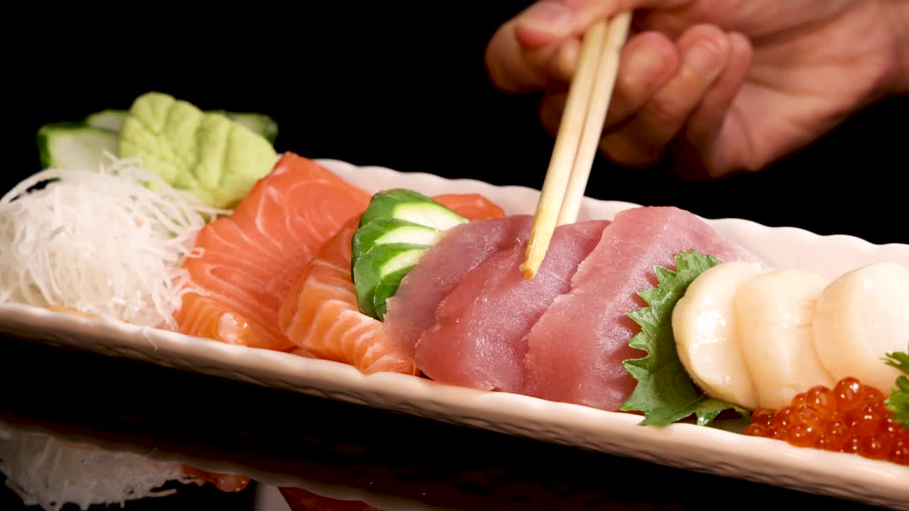 Hand uses chopsticks to select slices of salmon and tuna sashimi from a neatly arranged sushi platter under bright, focused lighting against a black background