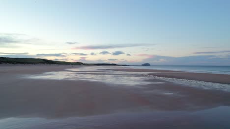 drone footage flying fast and low over a long sandy beach towards and island as a soft pink sunset reflects in tidal pools and the tide gently laps the shore of the beach