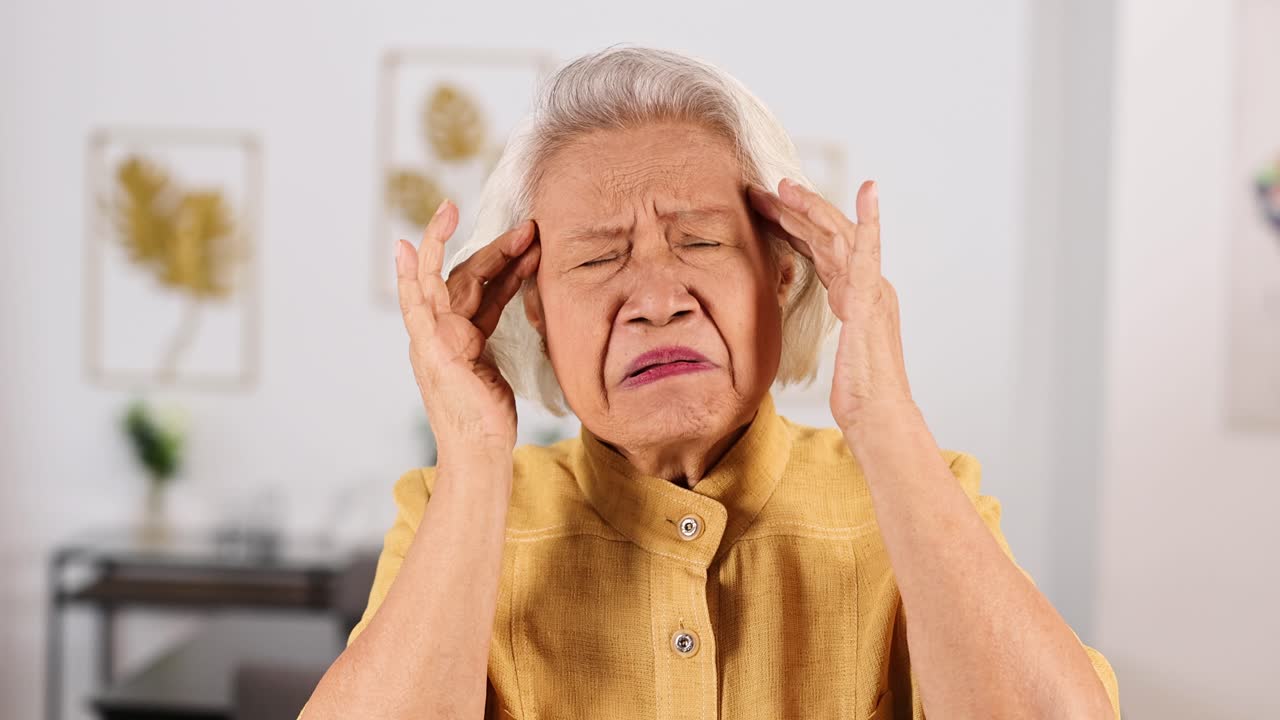 Senior Asian woman in yellow blouse holding head in pain, suffering headache at home interior