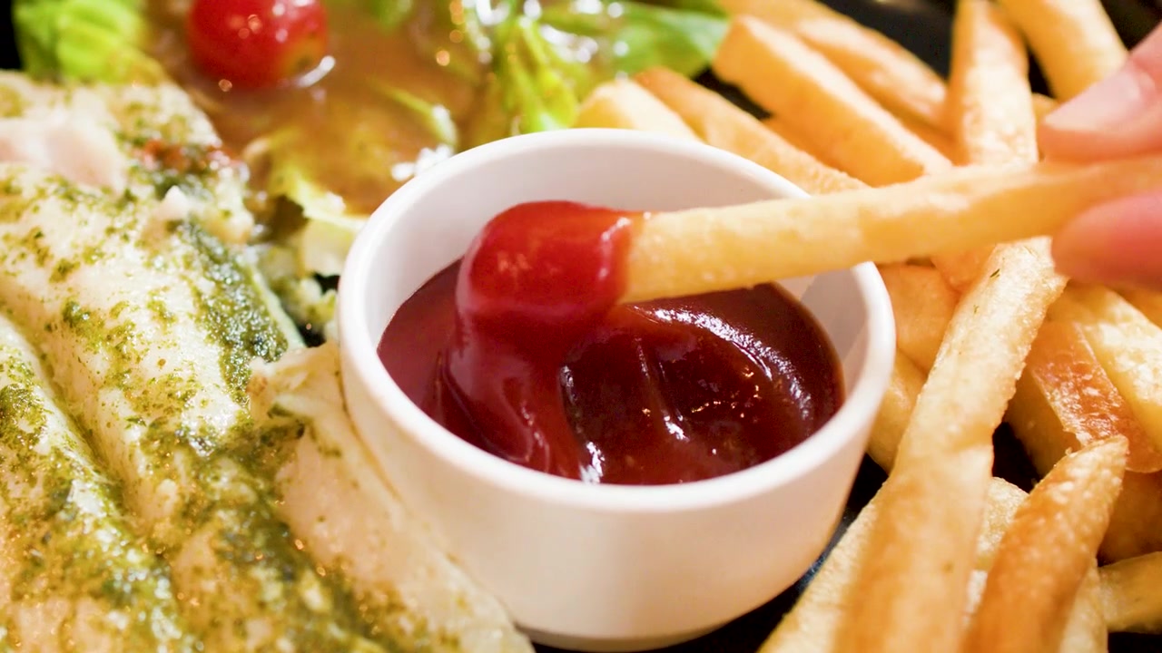 Eine goldene Pommes Frites taucht in roten Ketchup neben gegrilltem Fisch und Pesto-Soße