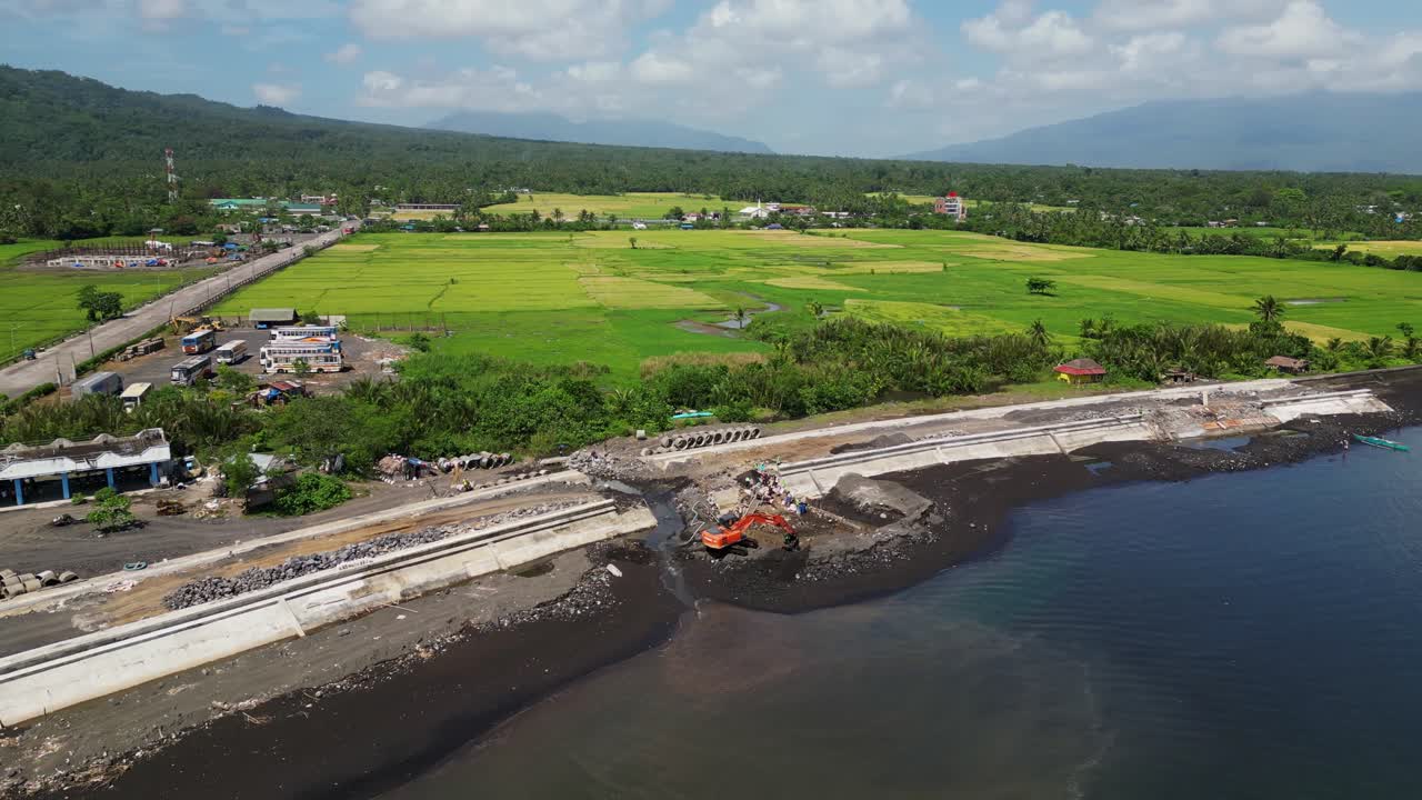 Aerial orbit of curved seaside road construction site and green fields in Malilipot, Albay near calm ocean water