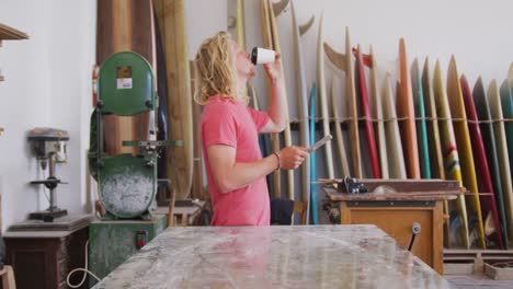 caucasian male surfboard makers using his smartphone and drinking coffee