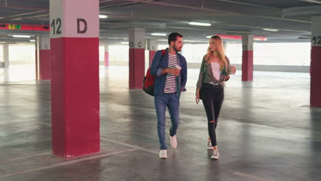 giovane uomo e donna caucasici ed eleganti che camminano in un parcheggio vuoto e parlano mentre bevono caffè