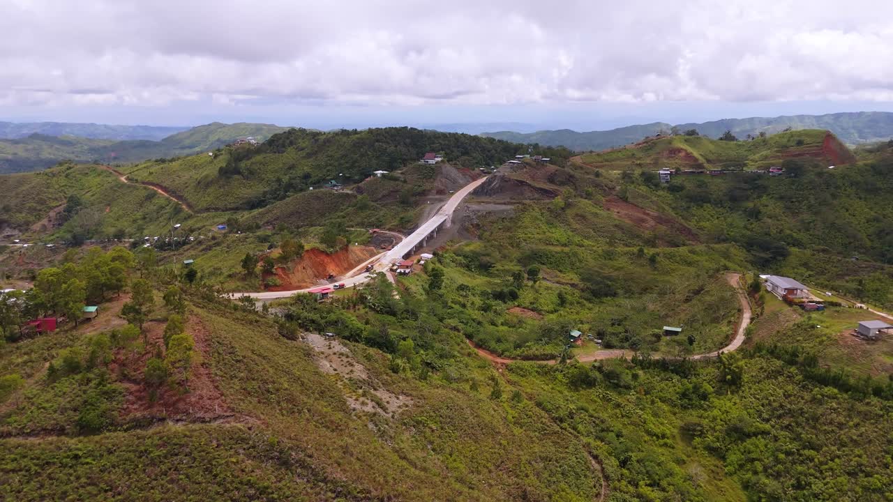 Drone footage of an elevated concrete bridge under construction in the mountains of the Philippines, surrounded by green hills, rural homes, and winding dirt roads under cloudy daylight