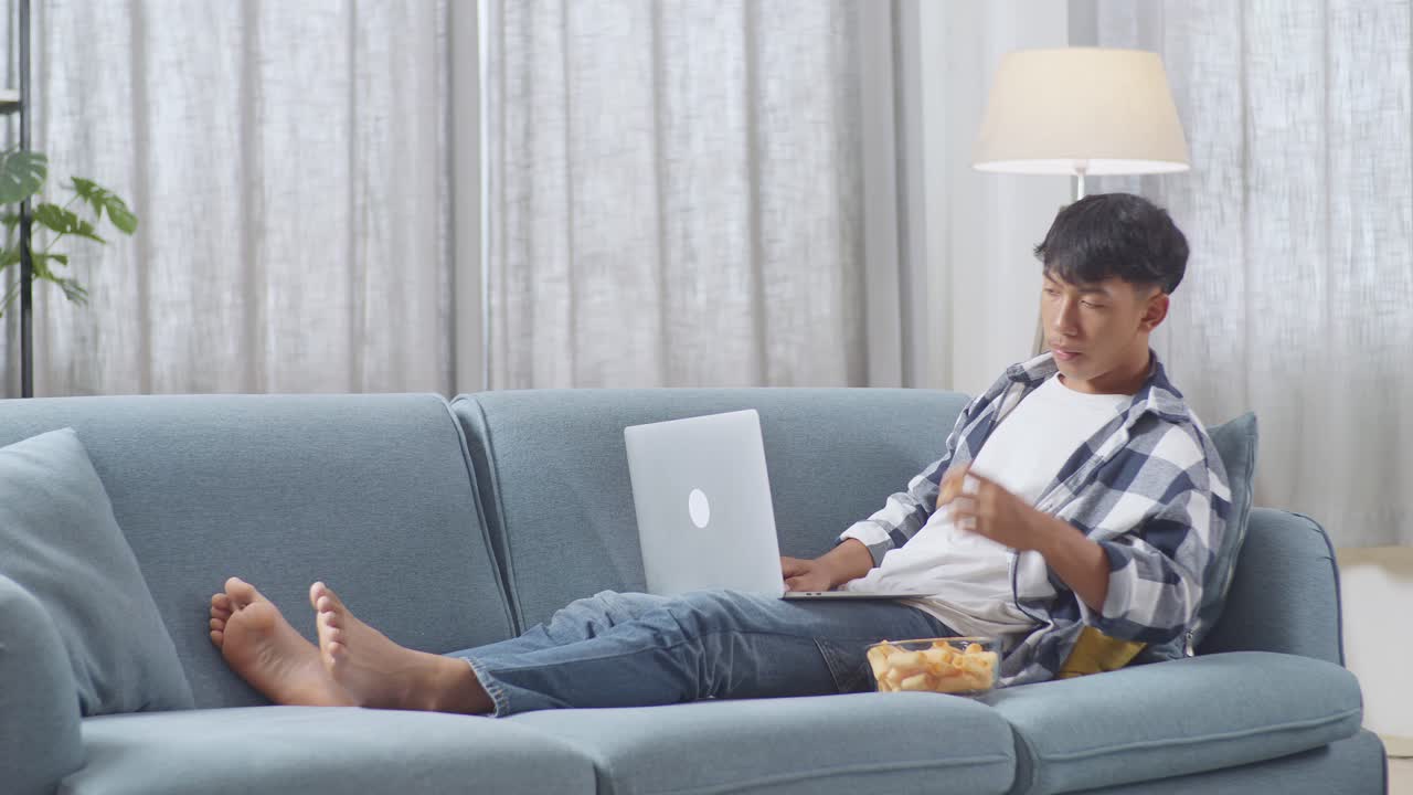 아시안 십대 소년의 몸이 가득 차 있으며, 집에서 거실에서 소파에 누워서 스을 먹고 노트북에 타이핑하고 있습니다.