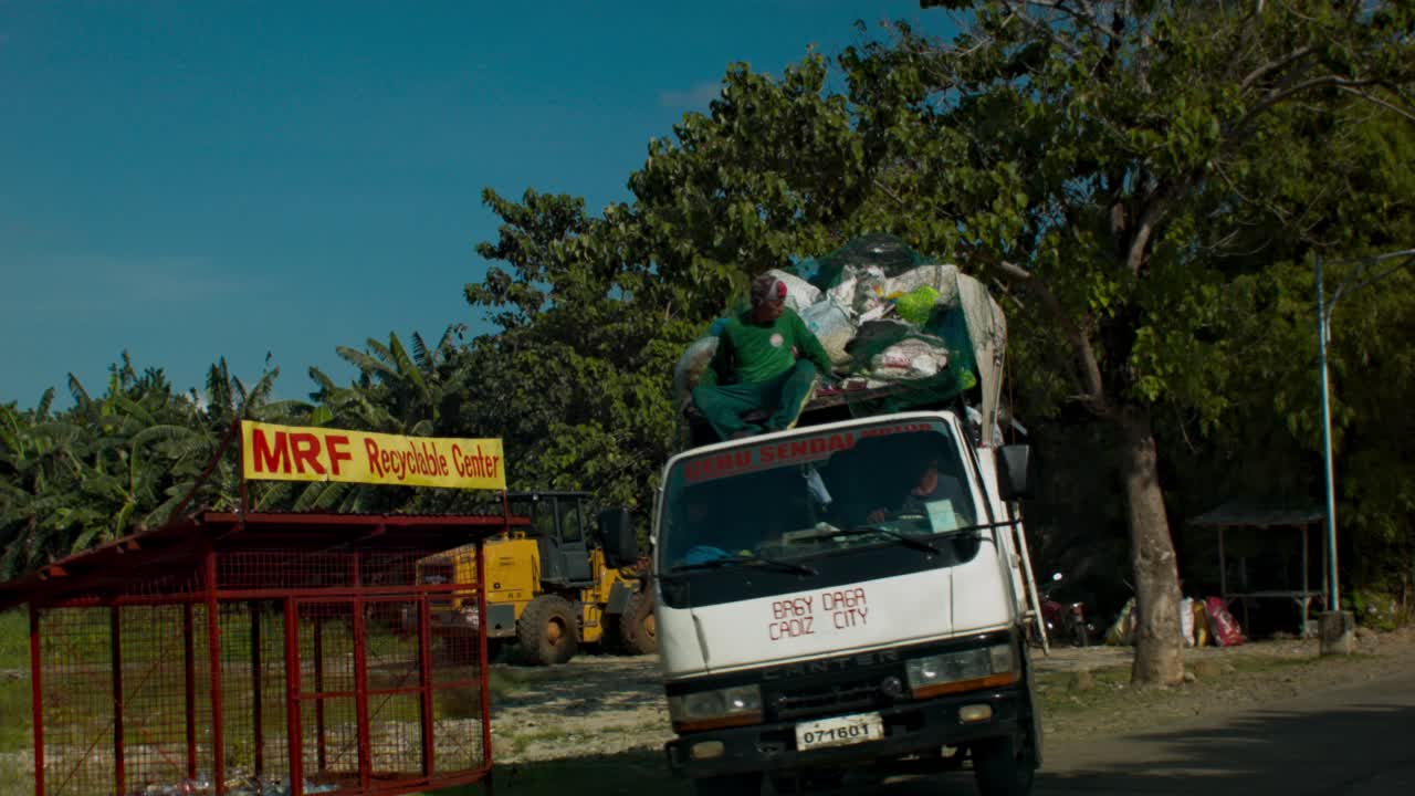 Garbage truck passes by. A garbage collector sits on the roof of the moving truck.