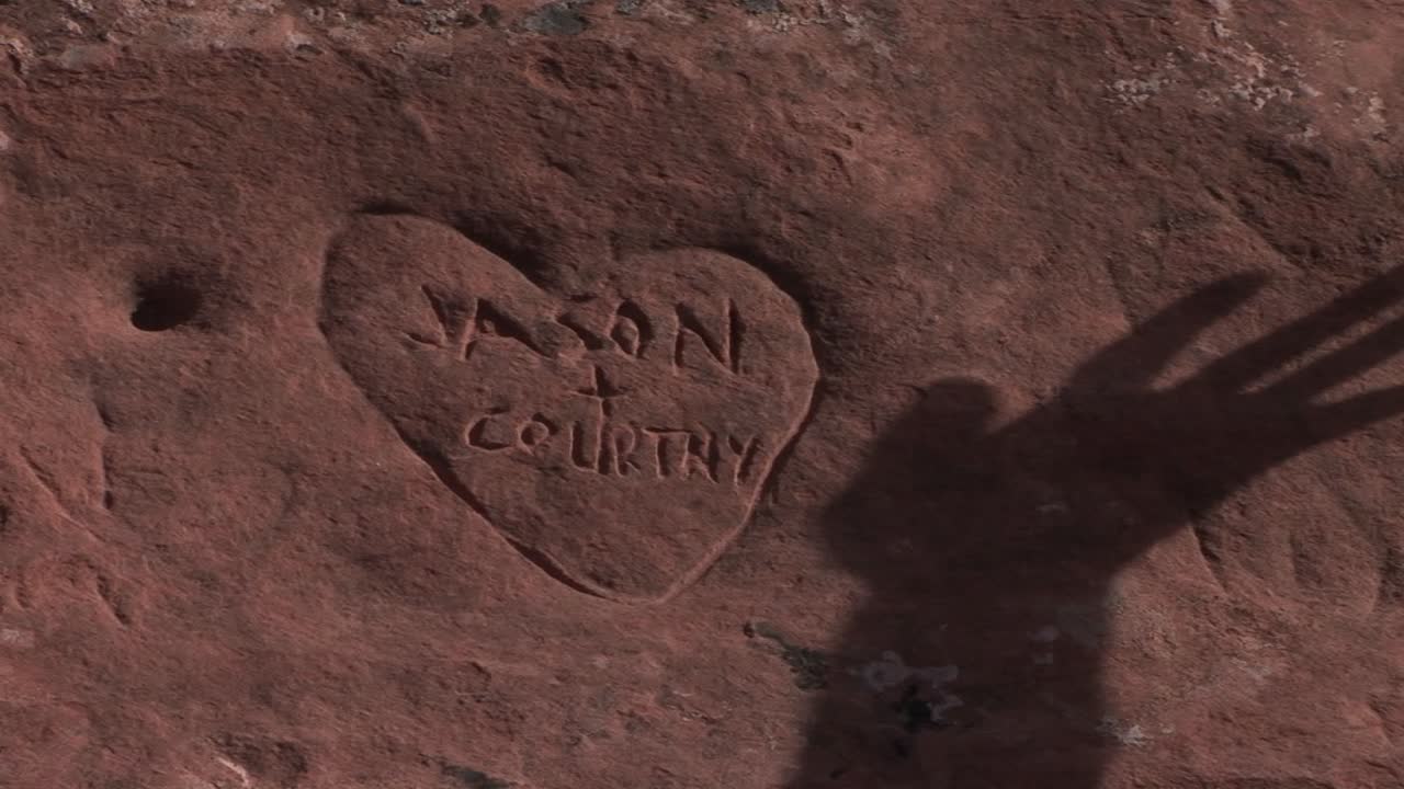 The shadow of a hand moves over a graffiti heart carved into sandstone