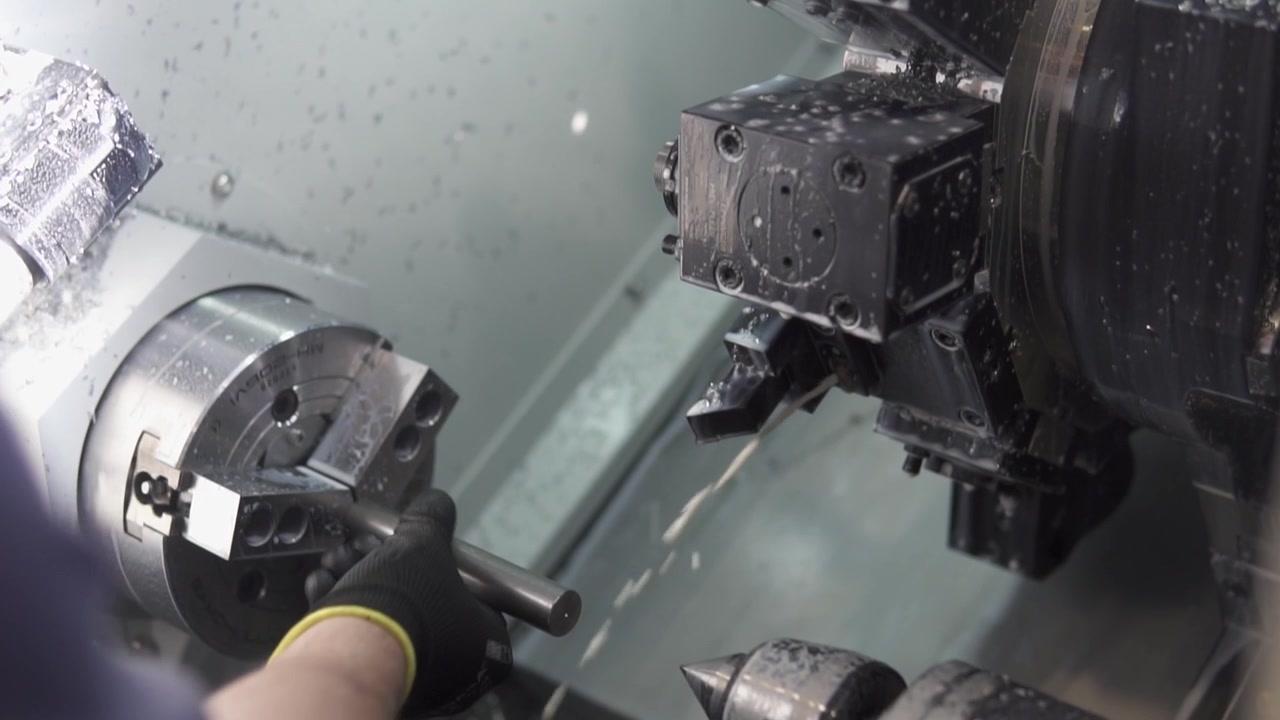 Technician in gloves operates CNC lathe, adjusting and monitoring metal machining in bright, controlled workshop. Clean, focused, industrial process.