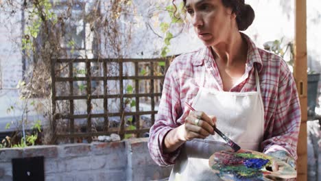 biracial female artist in apron painting on canvas in sunny garden, slow motion