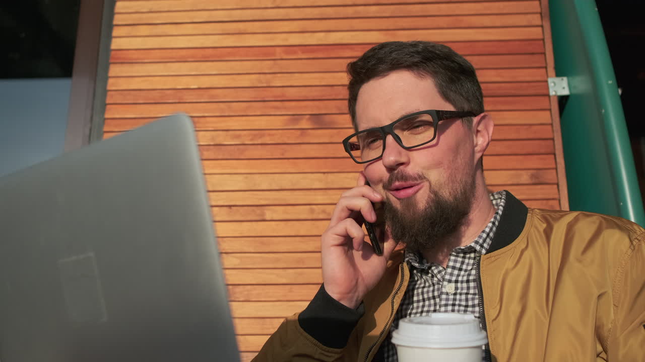 hombre trabajando al aire libre en una computadora portátil y hablando por teléfono mientras bebe café
