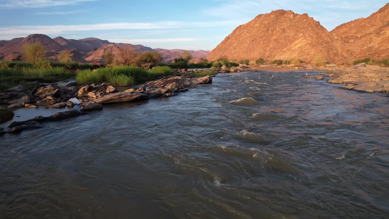 Smooth low-level drone flight over a rocky river with gentle rapids, ascending near the end to reveal stunning mountains. Perfect for nature, adventure, and landscape footage