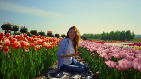 Young-woman-sitting-in-blooming-flower-garden-with-mobile-phone-in-sunny-day.