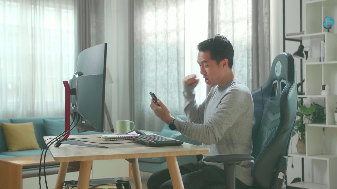 Asian Man In Long Sleeved T-Shirt And Black Pants Looking At Mobile Phone And Being Happy Winning The Project Bid While Using Desktop Computer For Working At Home.