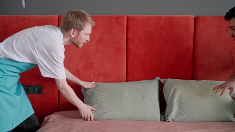 Side-view-of-a-blond-guy-with-a-beard-a-cleaner-in-a-white-T-shirt-and-a-blue-apron-together-with-his-colleague-a-brunette-guy-makes-the-bed-neatly-in-the-bedroom-cleaning-company