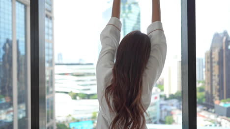 a young woman in a bathrobe and her back to the camera stretches while looking out the window at an urban skyline