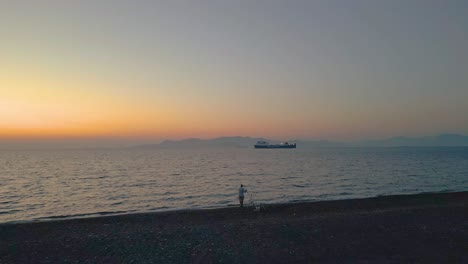 Hombre-Pescando-En-La-Orilla-Con-Un-Carguero-Navegando-En-El-Océano-Durante-El-Amanecer-En-Kos,-Grecia