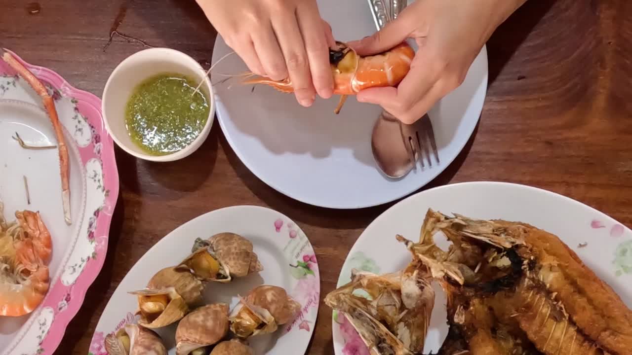 Close-up of hands peeling shrimp with sauce and seafood on a wooden table.