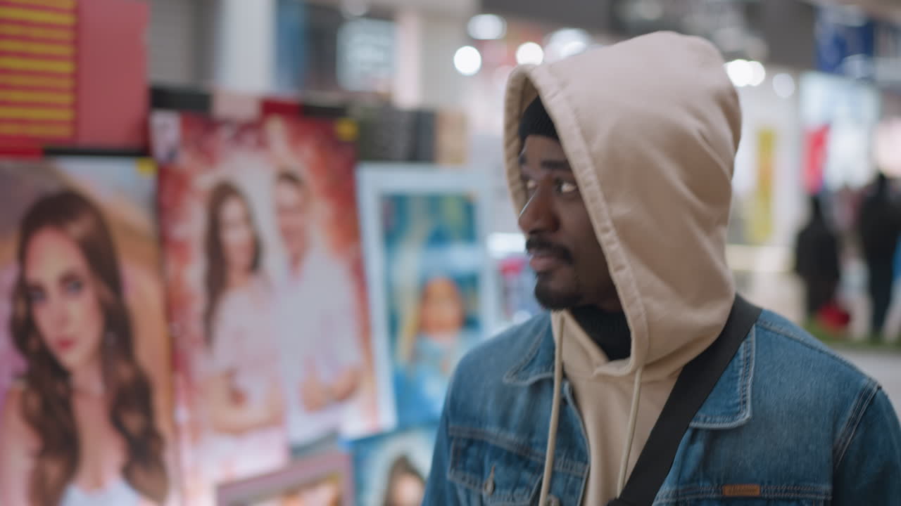 close shot of male walking through busy indoor mall wearing beige hoodie denim jacket and beanie under hood backpack strap visible against blurred store lights and modern interior