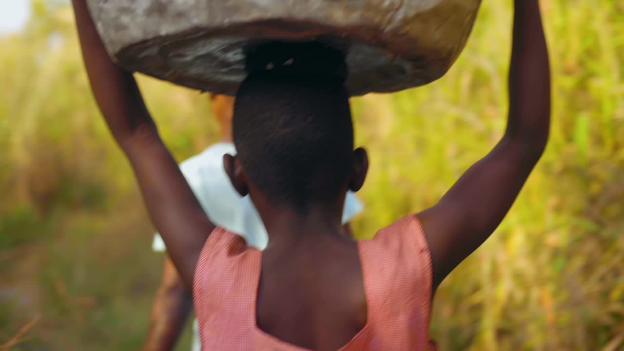 Group Of Local Young African Women And Kids Are Carrying Big Heavy Buckets  Of Drinking Water And Bringing The Water On Their Heads Towards The Village  Walking In Bush Field - Close