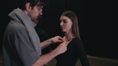 Confident-brunette-man-in-a-gray-T-shirt-and-scarf-the-stage-director-attaches-a-small-microphone-to-the-black-suit-of-an-actress-before-going-on-stage-during-a-performance-in-the-theater