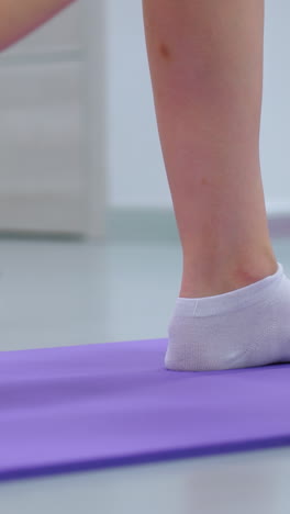 vista cercana de la pierna de una mujer con calcetines levantando un peso en una alfombra púrpura, mostrando un movimiento de ejercicio dinámico con otra persona parcialmente visible en el fondo