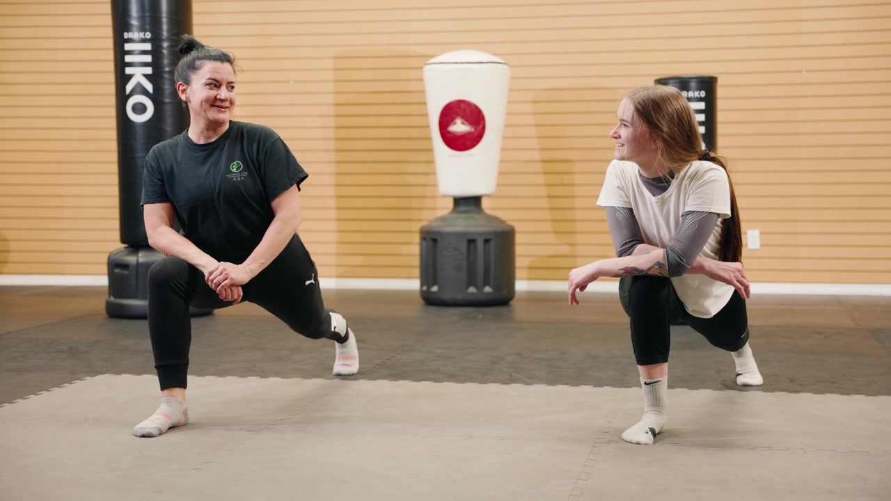 Bidikan Jarak Menengah: Kelompok Ibu dan Anak Perempuan Paruh Baya Melakukan Lunges Atletis dan Peregangan di dalam Pusat Kebugaran/Gym Seni Bela Diri Taekwondo, dengan Karung Tinju di Latar Belakang, Berbicara, Tersenyum, dan Membangun Ikatan
