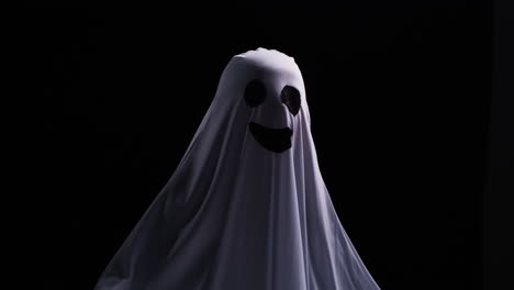 유령 의상을 입은 아이가 할로윈에 트릭 또는 치료를 하고 검은 스튜디오 배경 앞에서 사람들을 겁내고 있습니다.