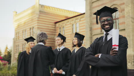 porträt eines afroamerikanischen lächelnden männlichen absolventen, der vor der universität posiert und seine hände kreuzt